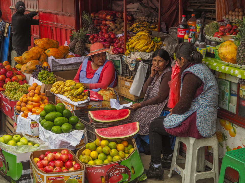 Trois femmes assises derrière un assortiment de fruits exposés sur le trottoir