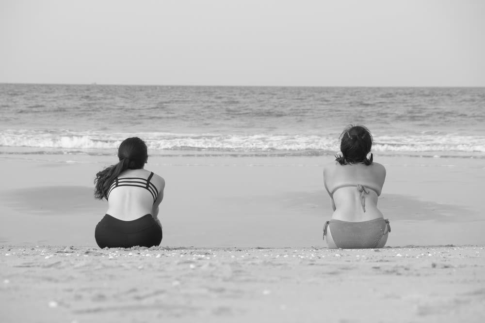 two women sitting on shore front of calm sea
