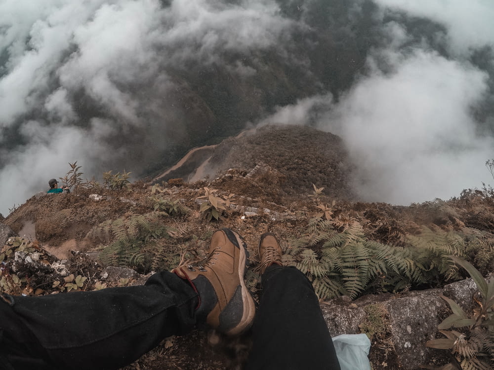 person in brown shoes and black pants sitting on edge of mountain