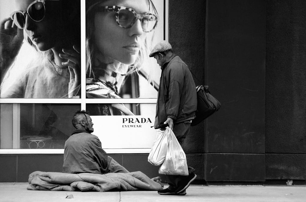 grayscale photo of man with shop bags walking past beggar siting on sidewalk