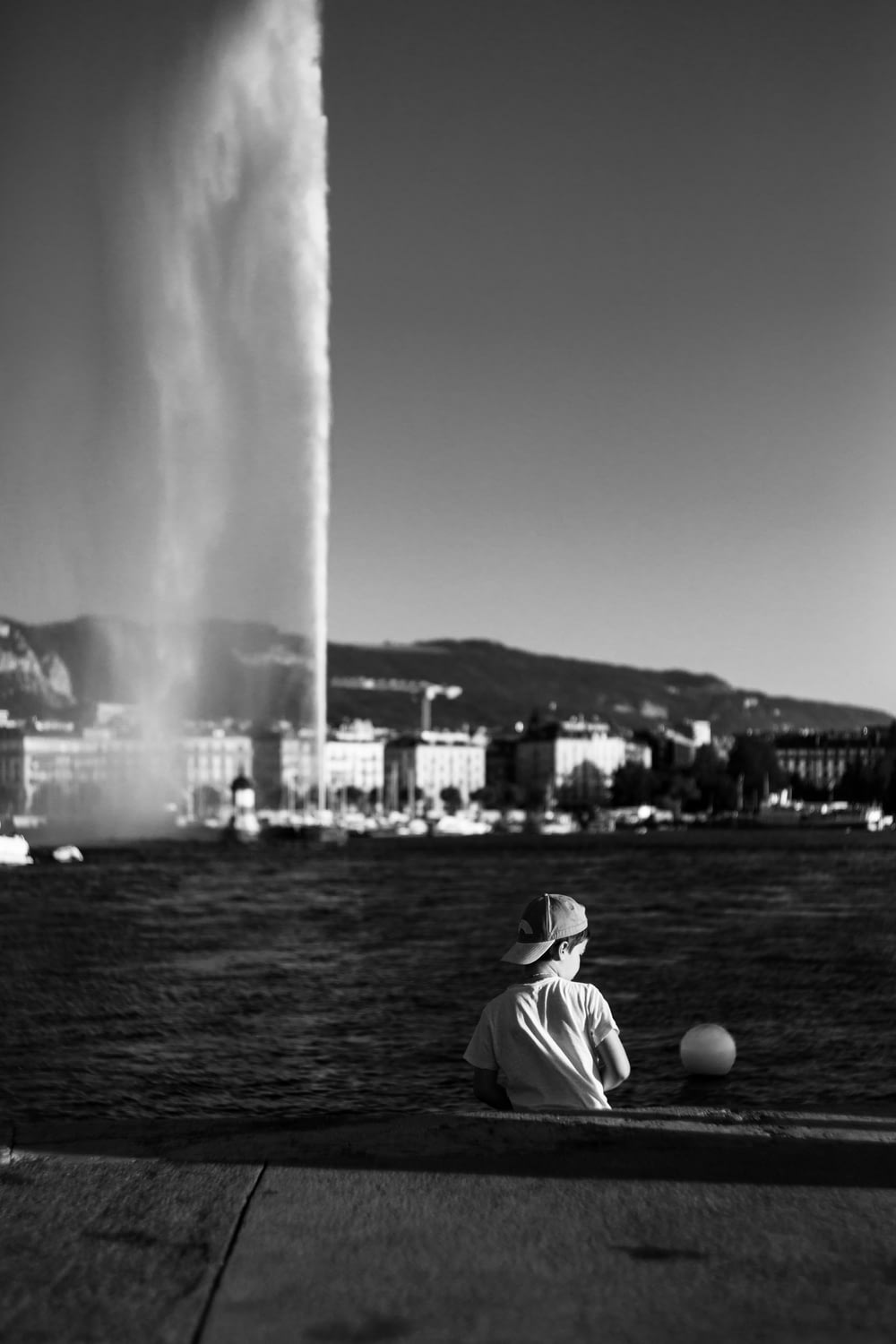 grayscale photography of boy sitting beside body of water