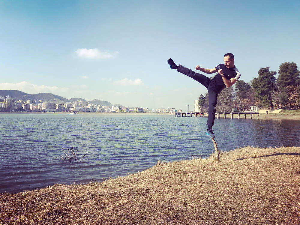 man standing on on leg on twig doing kicking pose beside body of water during day