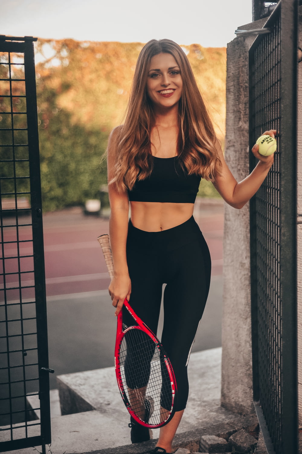 smiling woman wearing black sports bra and black leggings holding red tennis racket and ball standing beside black metal gate