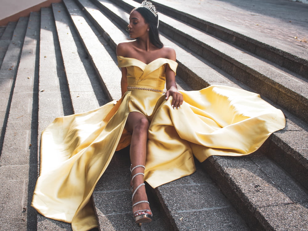 woman wearing yellow off-shoulder dress with silver tiara sitting on gray concrete stairs