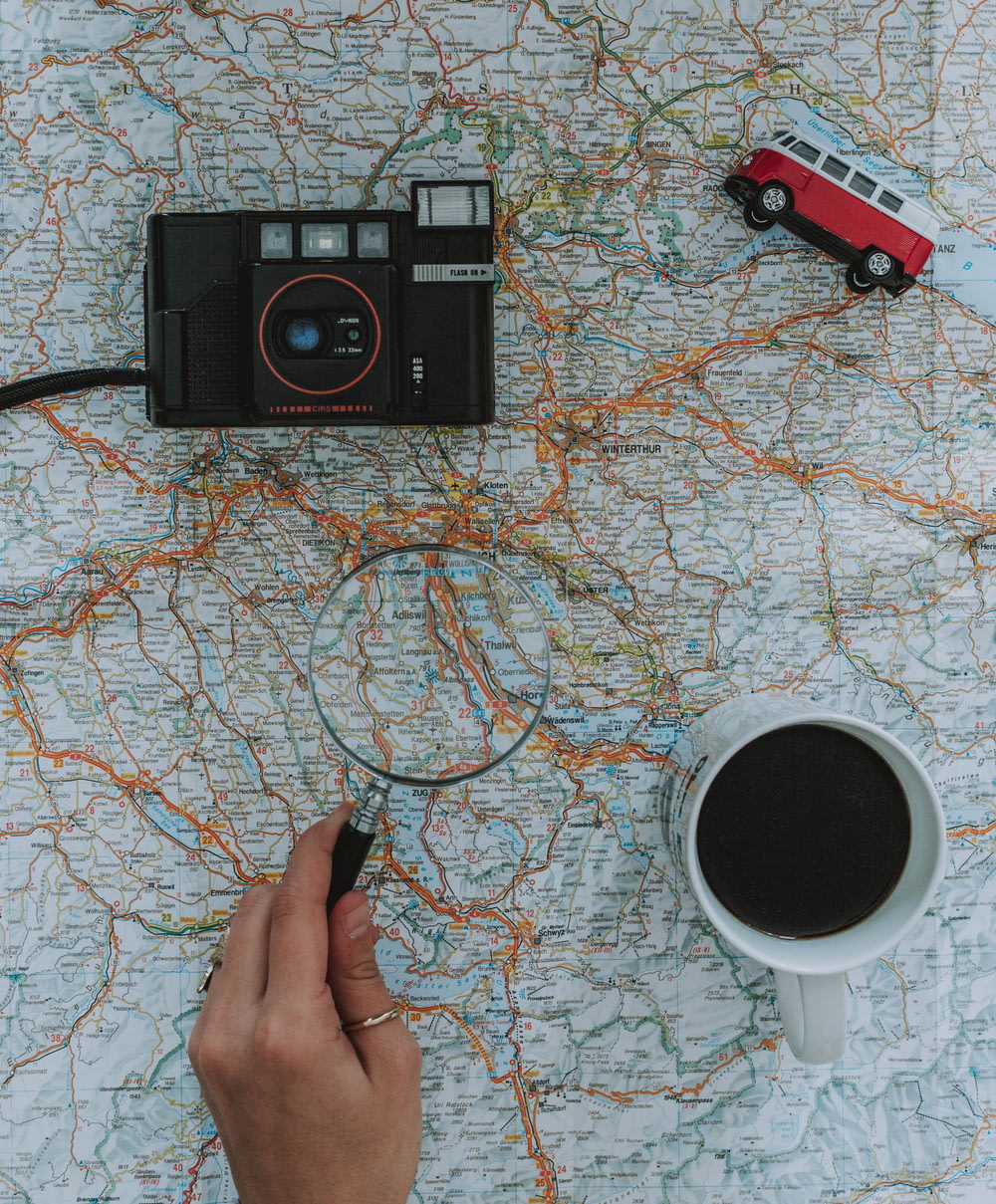 person holding magnifying glass beside black film camera white ceramic mug filled with black liquid near vehicle toy scale model