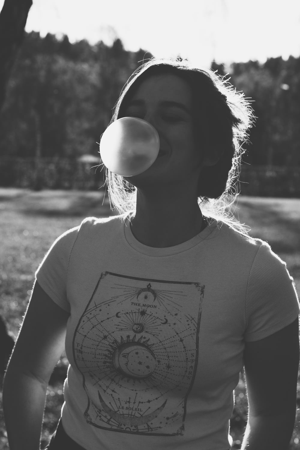grayscale photography of woman wearing crew-neck t-shirt standing while making bubble from chewing gum