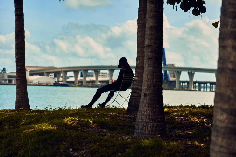 person wearing cap sitting on chair under tree beside body of water during daytime