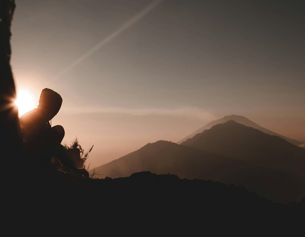 silhouette of person wearing hoodie sitting on mountain while looking at sunset scenery