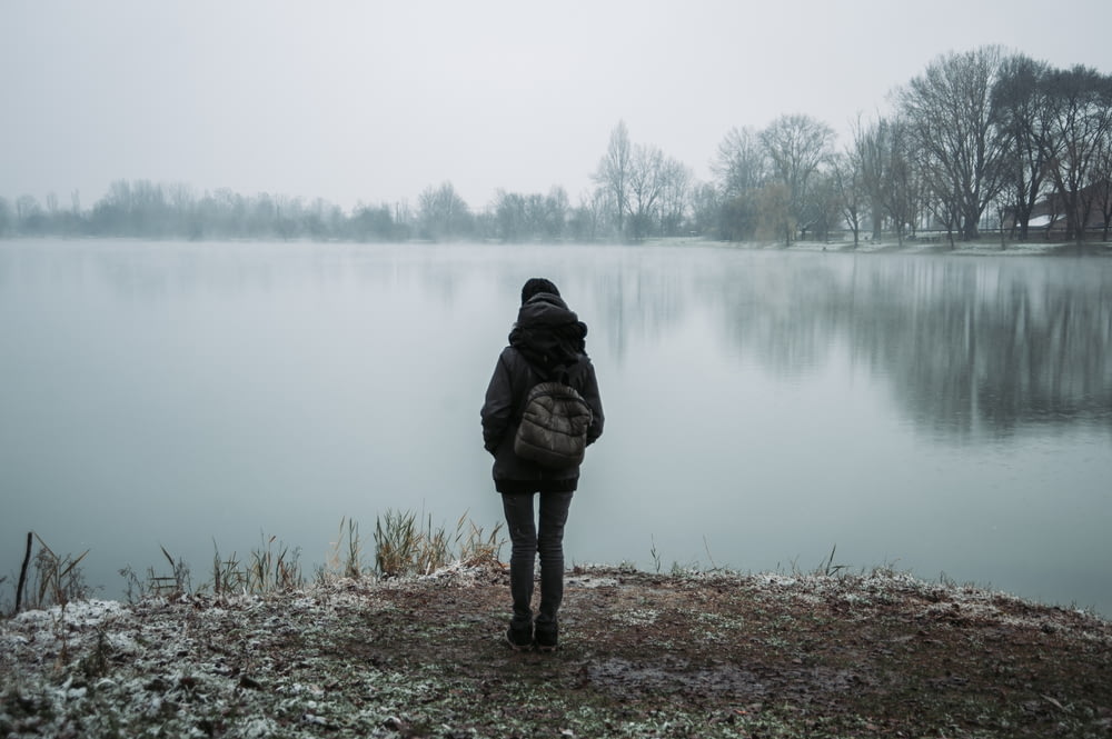person wearing black hooded jacket standing and facing on body of water near trees in foggy day