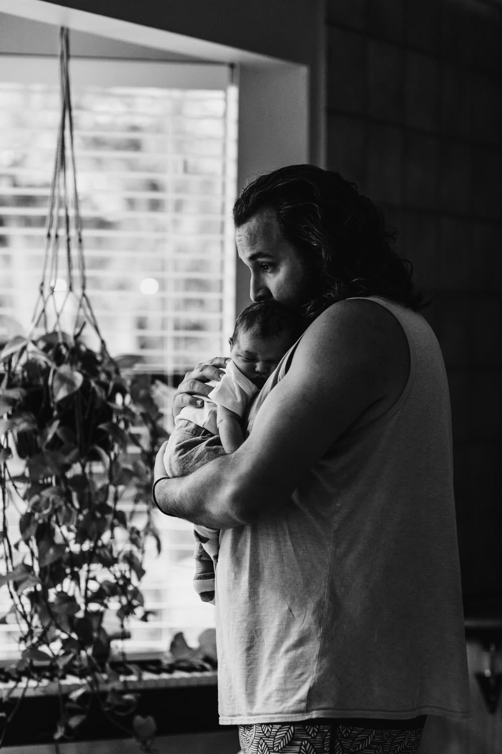 grayscale photography of man wearing tank top carrying newborn baby while standing near window