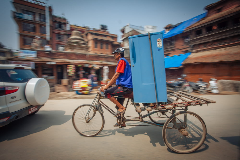 man riding adult trike with blue single-door refrigerator on road near car during day