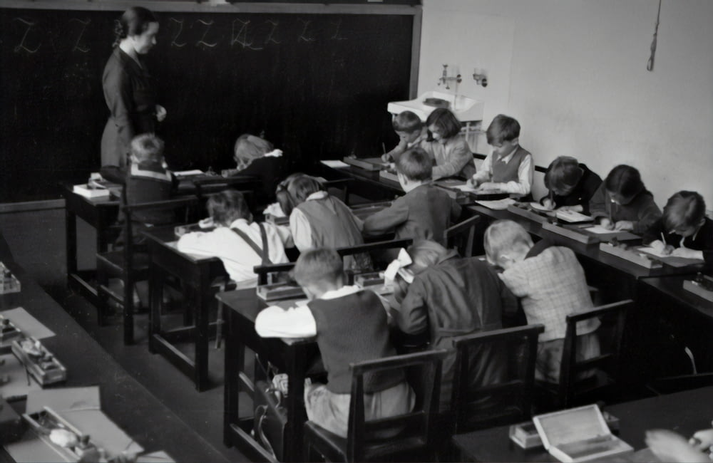 fotografia em escala de cinza do professor em pé perto do quadro-negro e das crianças sentadas em cadeiras