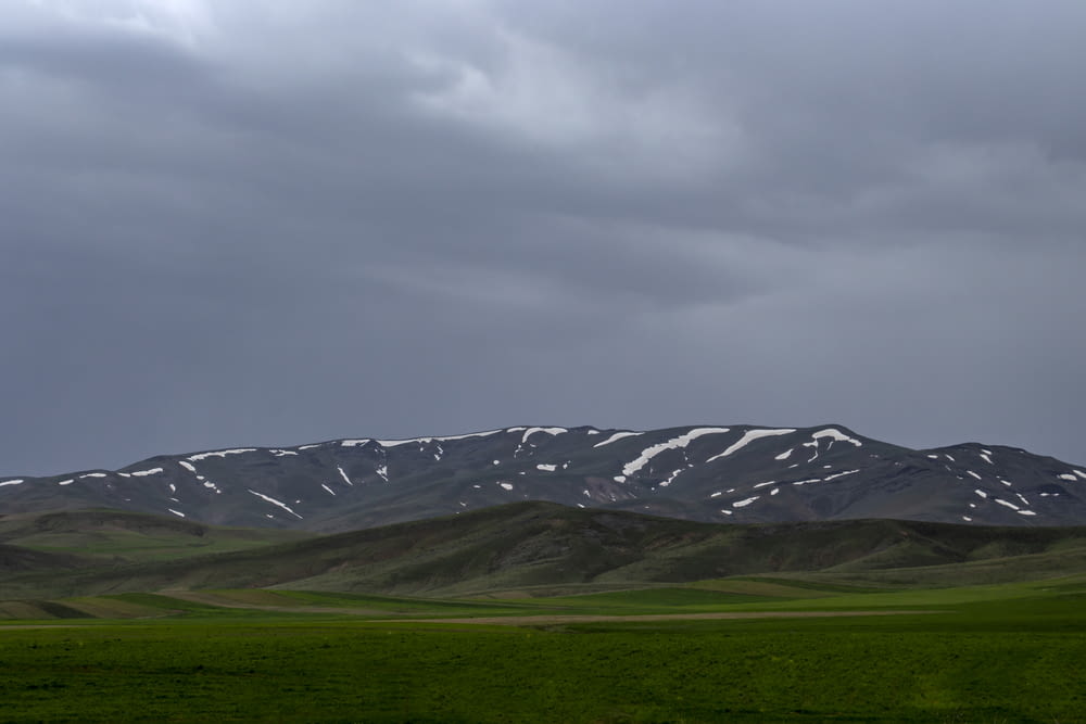 green grass field near snow covered mountain under cloudy sky during daytime