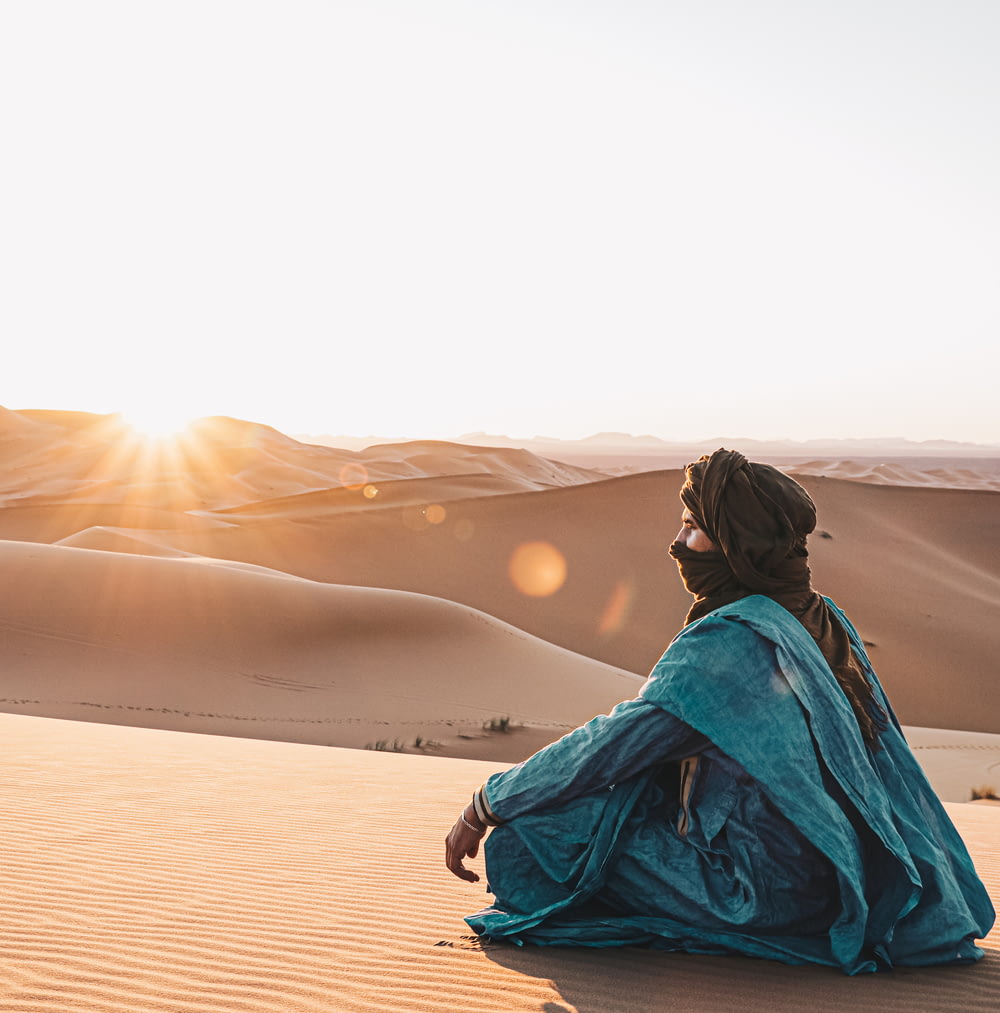 Frau in blauem Kapuzenpulli sitzt bei Sonnenuntergang auf Sand
