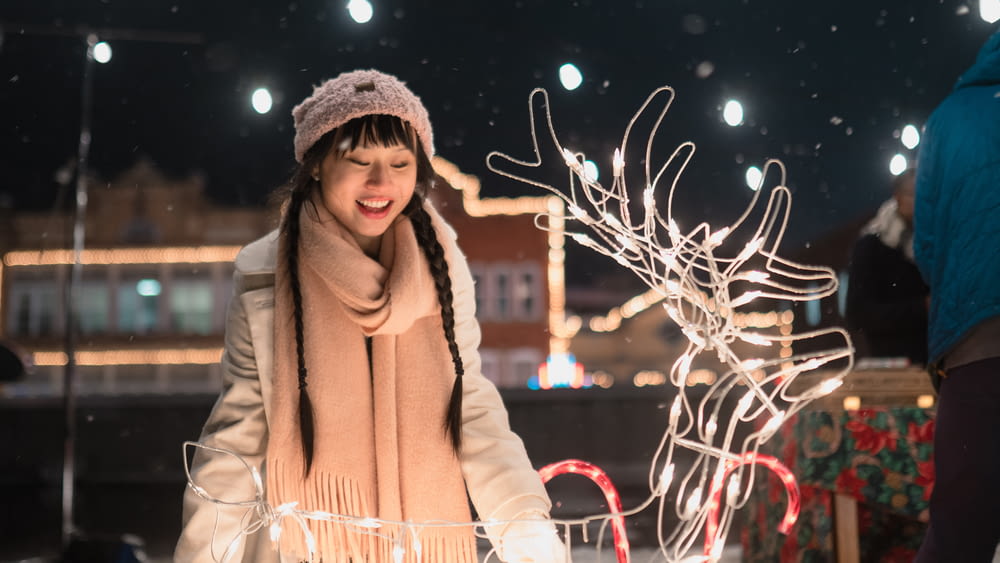 woman in brown coat standing near white string lights during daytime