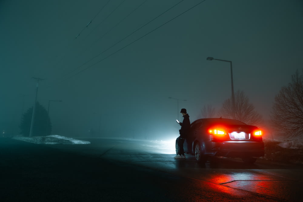 검은 재킷과 검은 바지를 입은 남자가 밤 시간에 빨간 차 옆에 서 있다
