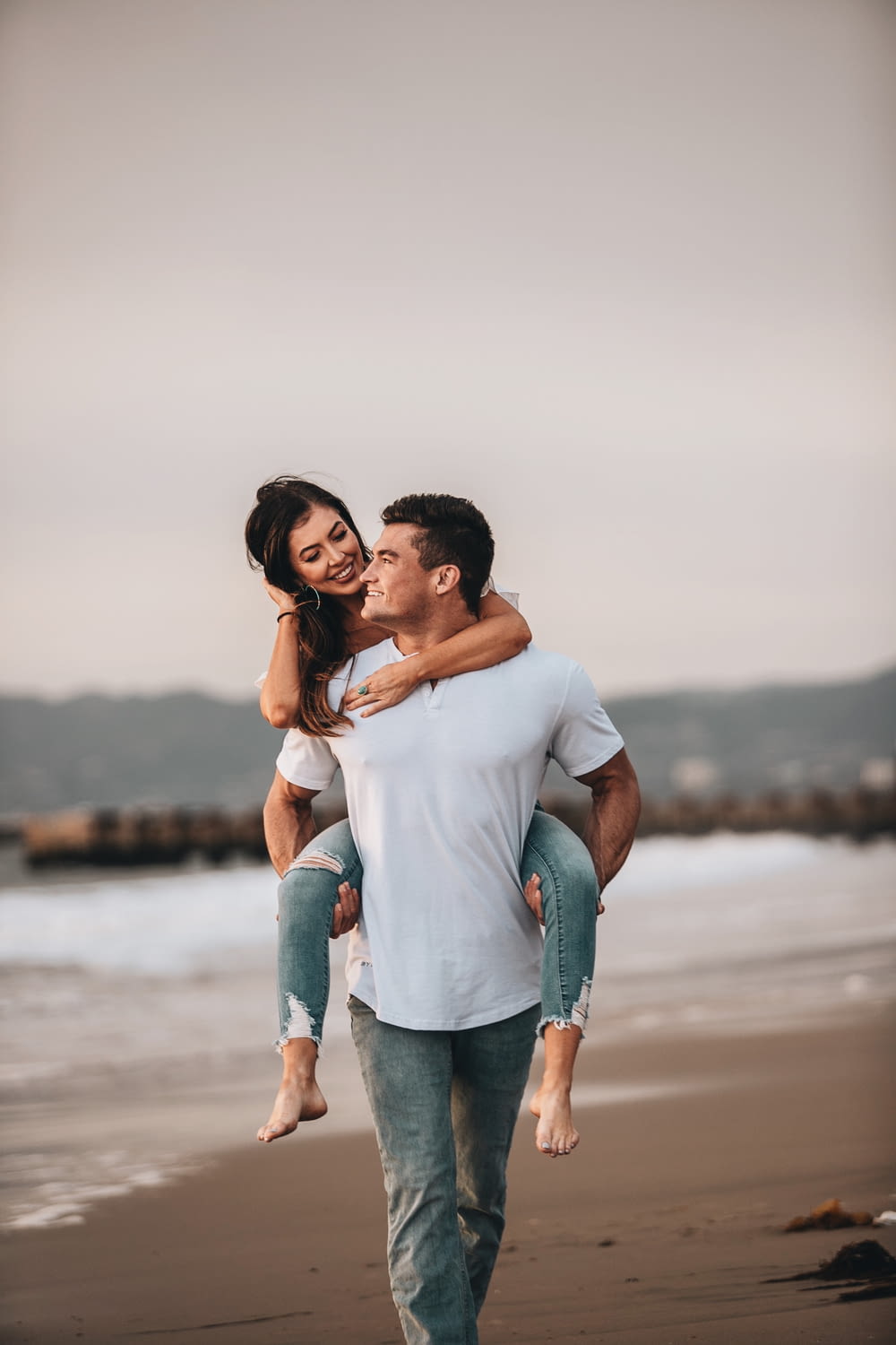 man in white crew neck t-shirt kissing woman in blue denim jeans on beach during