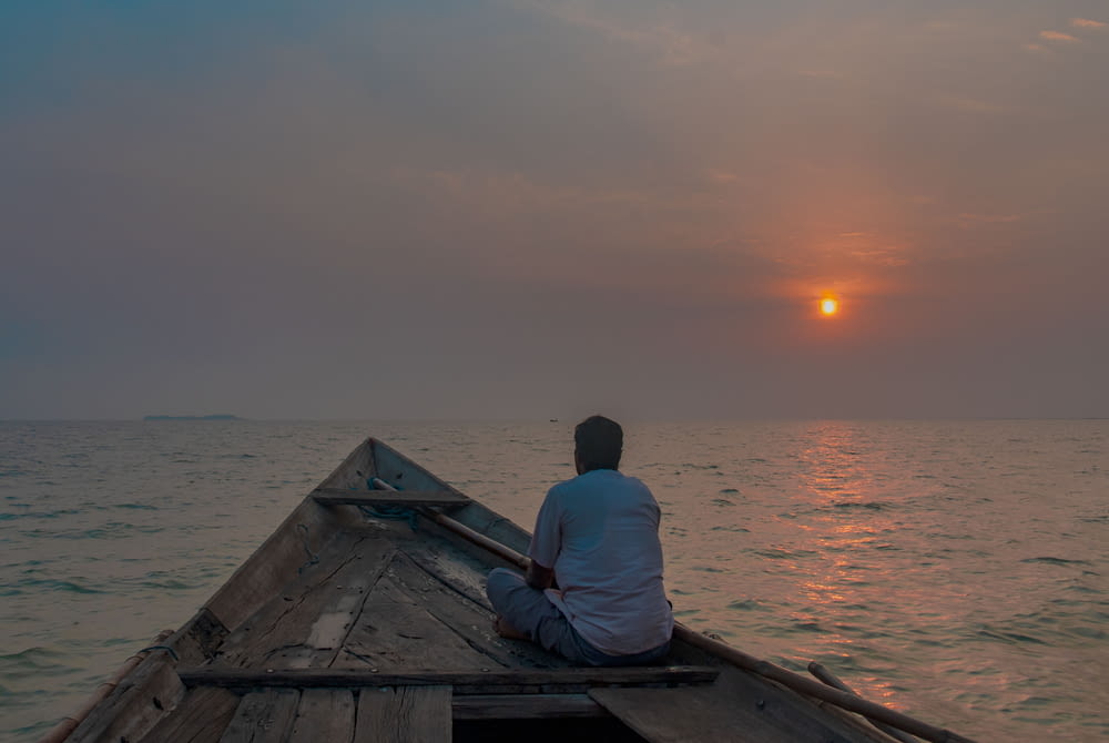 man in white shirt sitting on brown wooden boat on sea during sunset