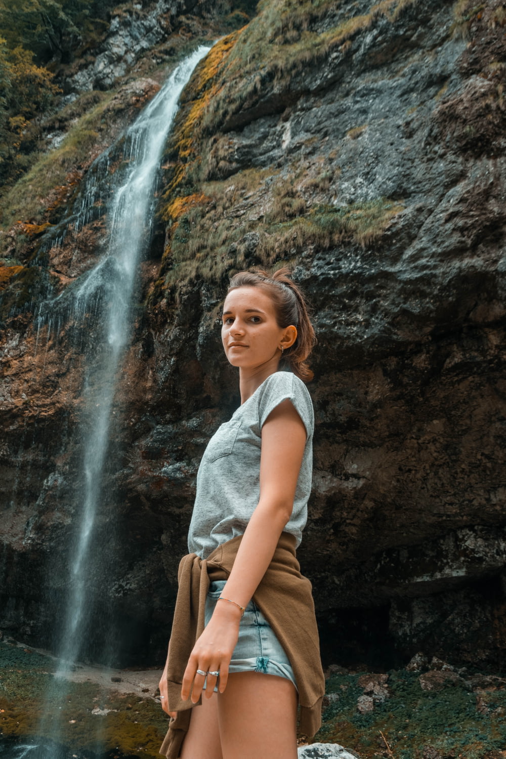 woman in white tank top and brown pants standing near waterfalls during daytime