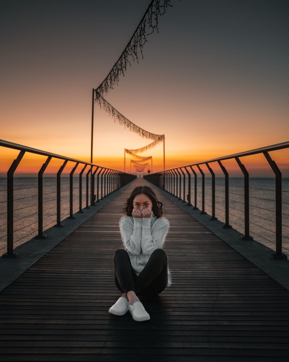 woman in white long sleeve shirt and white pants sitting on wooden dock during sunset