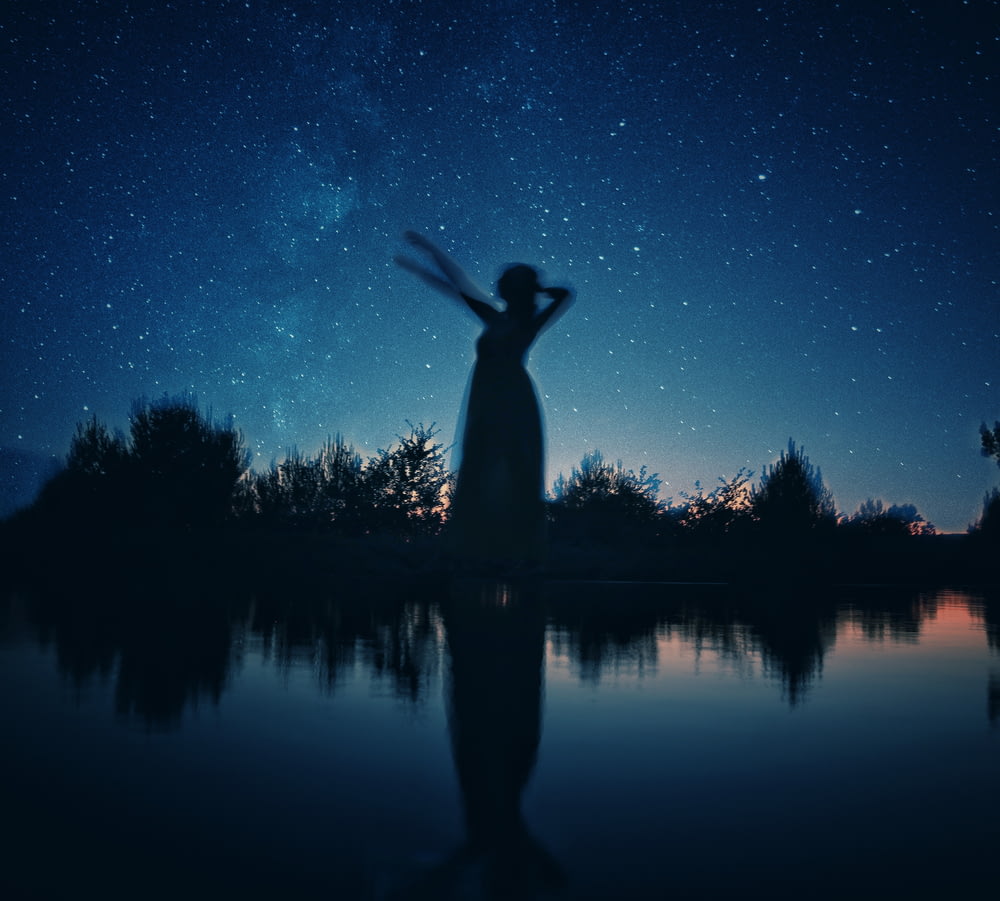 silhouette of woman standing on rock near body of water during night time