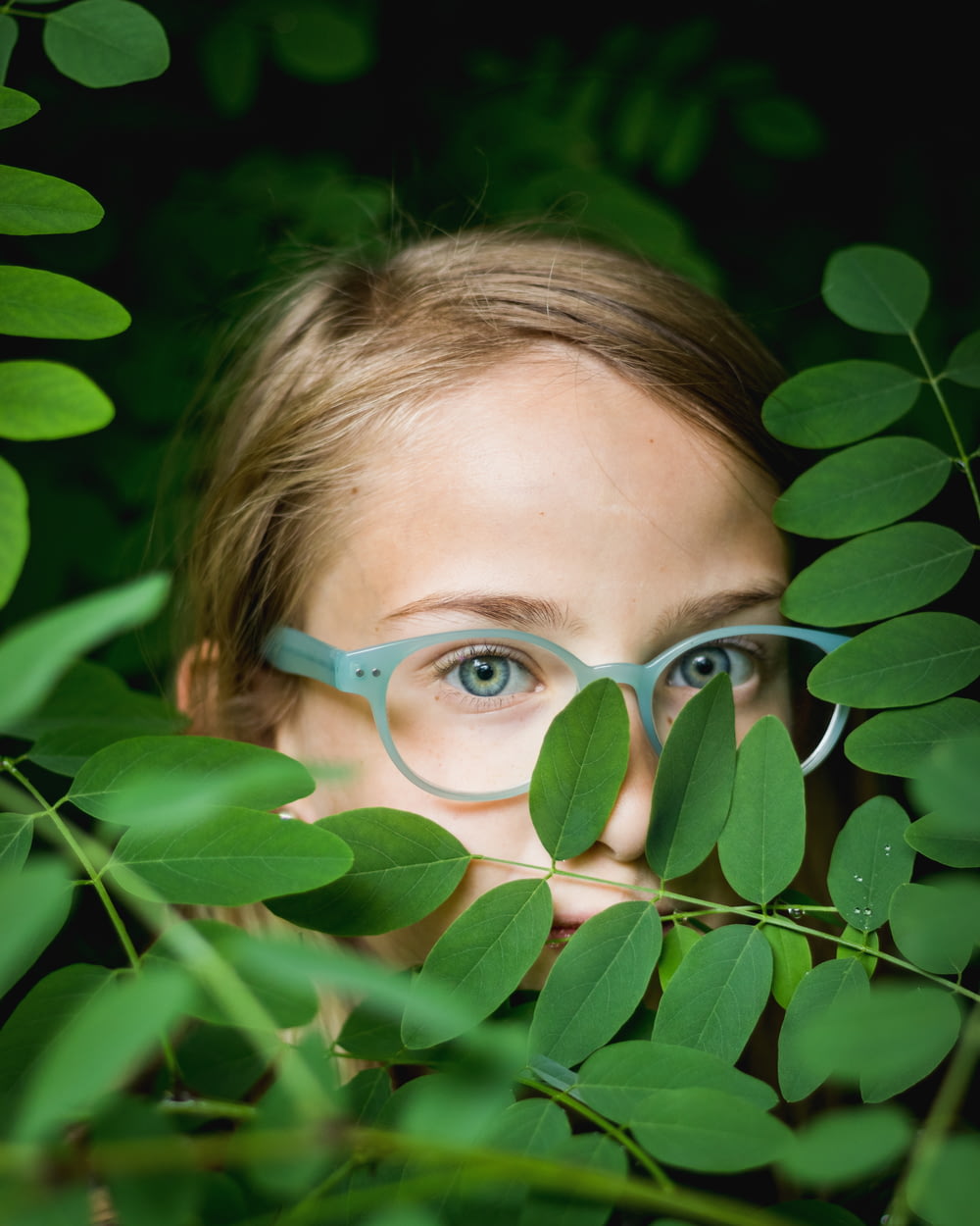 chica con gafas de marco azul que se esconde detrás de las hojas verdes
