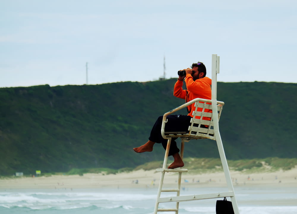 uomo in camicia arancione che si siede sulla sedia pieghevole di legno bianca sulla spiaggia durante il giorno