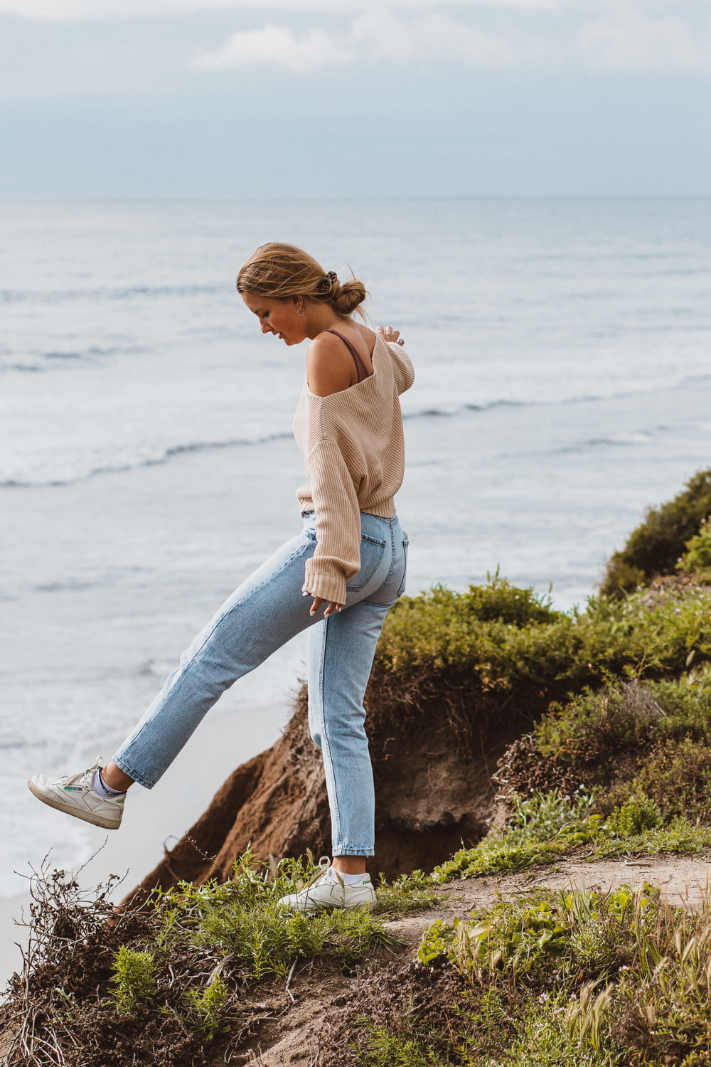 woman in brown long sleeve shirt and blue denim jeans sitting on rock near sea during