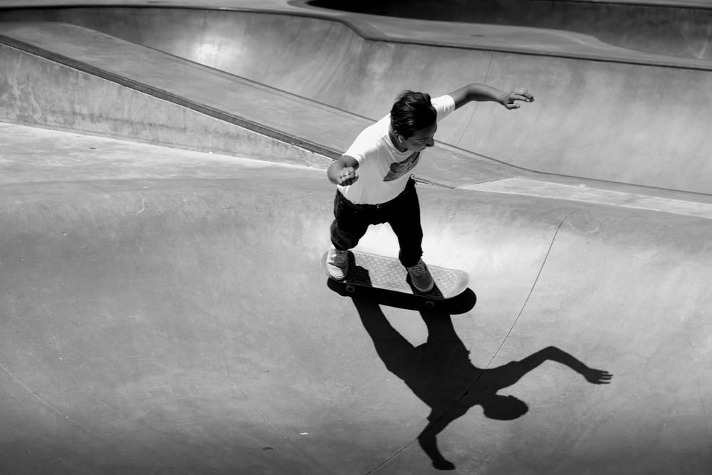 Photo en niveaux de gris d’un homme en t-shirt blanc et pantalon noir faisant des cascades de skateboard