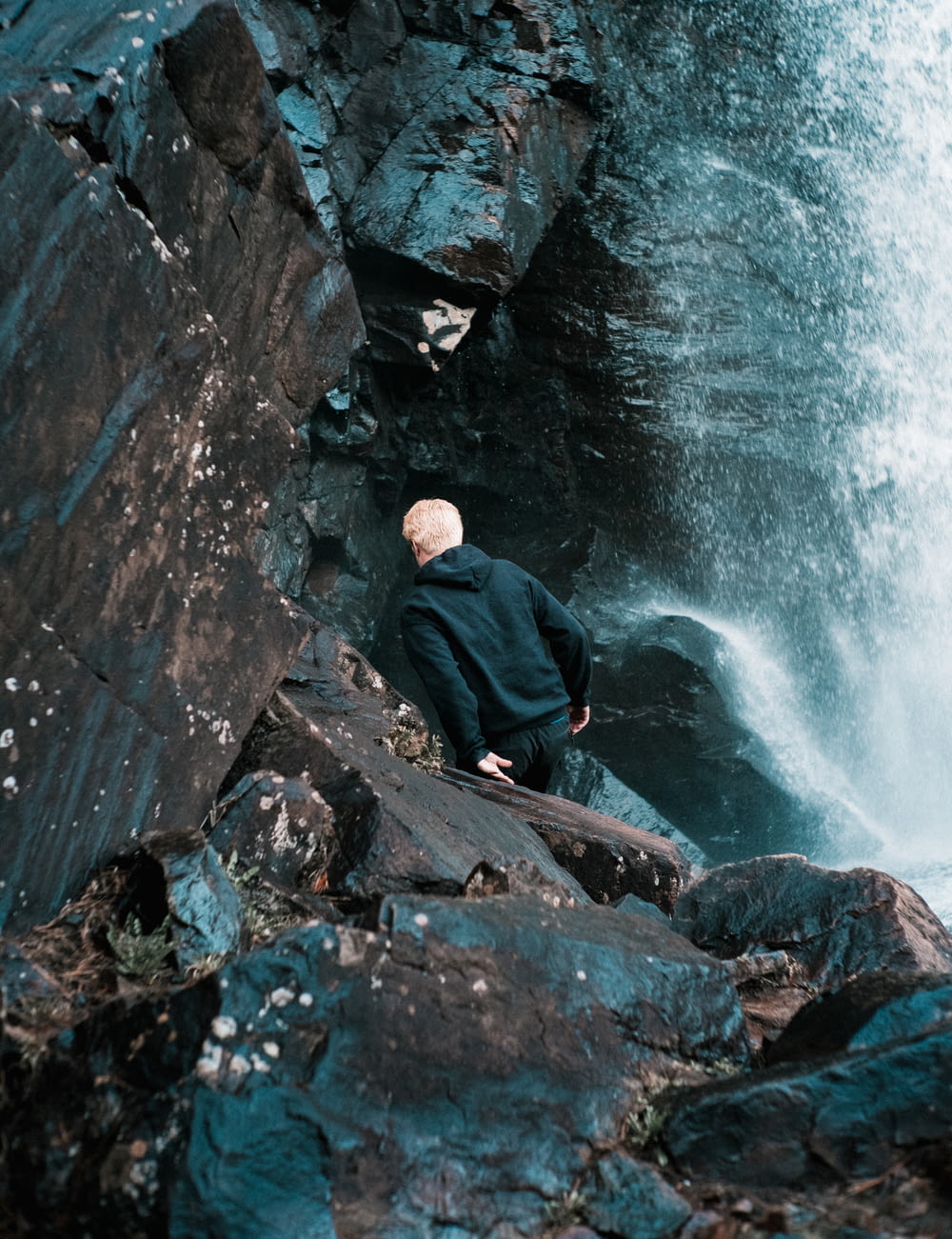 man in black jacket sitting on rock near water falls during daytime