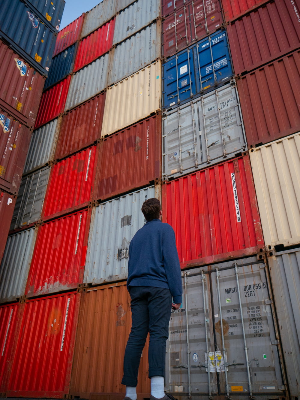 uomo in camicia blu a maniche lunghe in piedi davanti ai container intermodali rossi e blu