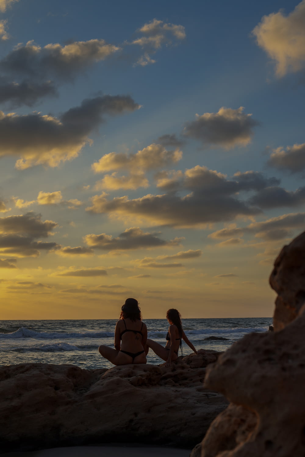 Frau im schwarzen Bikini sitzt bei Sonnenuntergang auf einem Felsen in der Nähe des Meeres