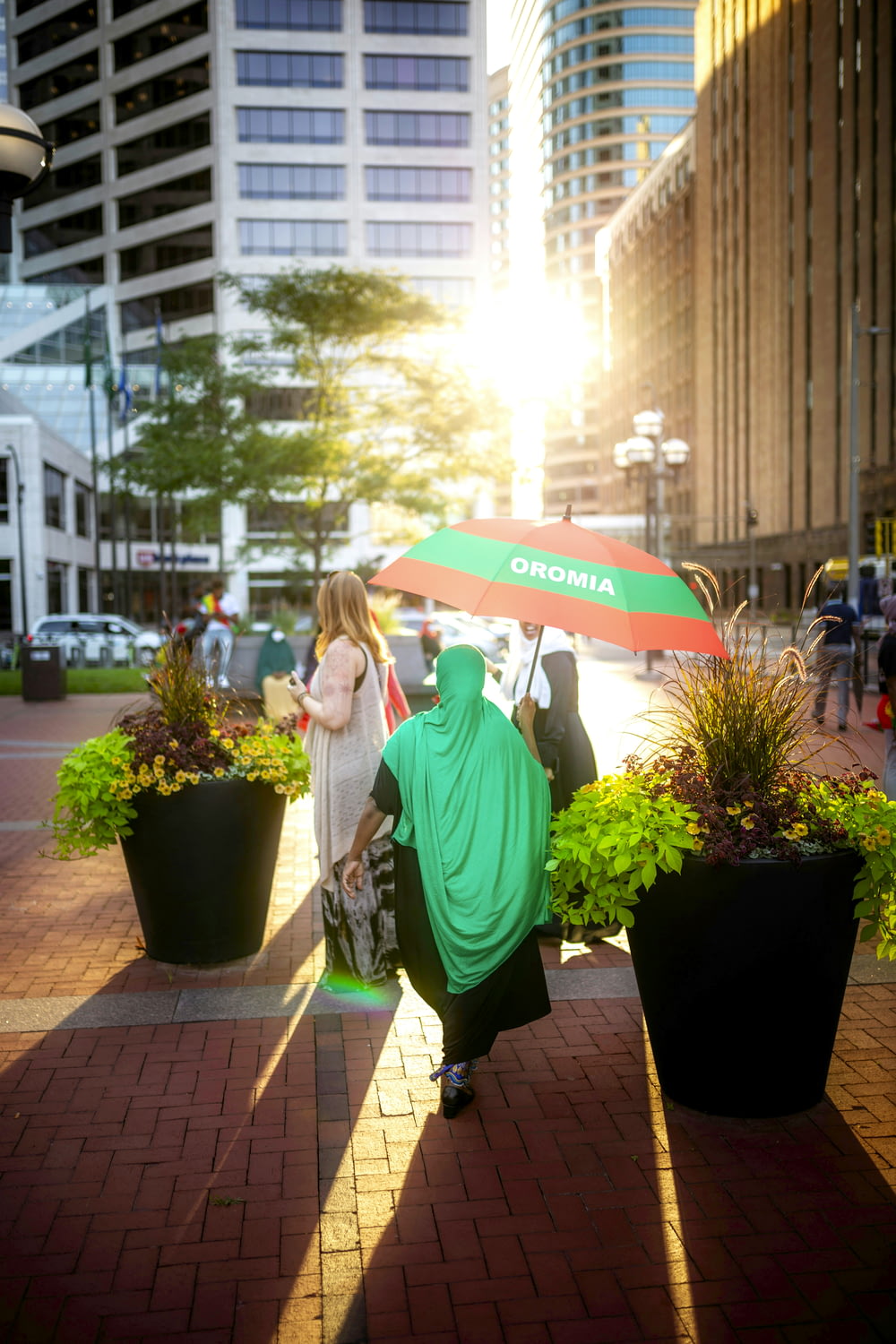 昼間、緑の鉢植えのそばに傘をさす緑のパーカーを着た女性