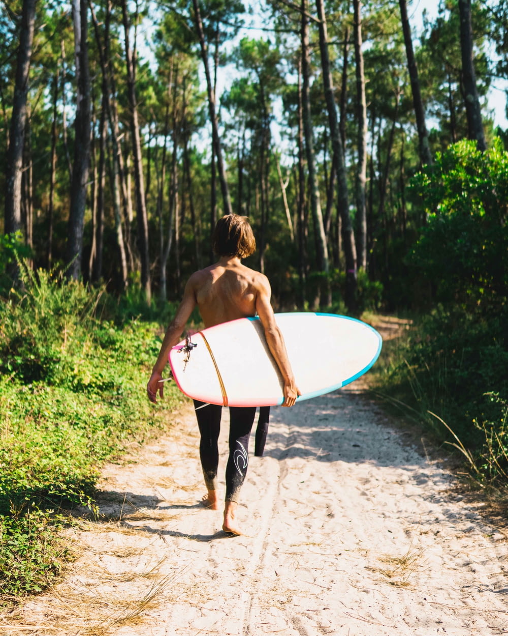 woman in black and white bikini holding white surfboard walking on dirt road during daytime