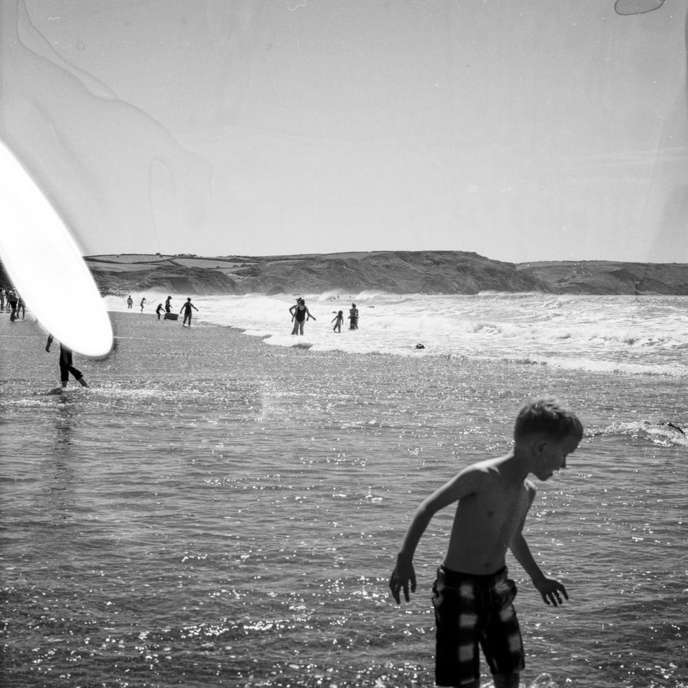 Foto en escala de grises de niño sosteniendo una tabla de surf en la playa