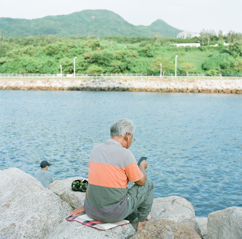 homem em camiseta laranja e calças cinzas sentado na rocha cinza perto do corpo de água