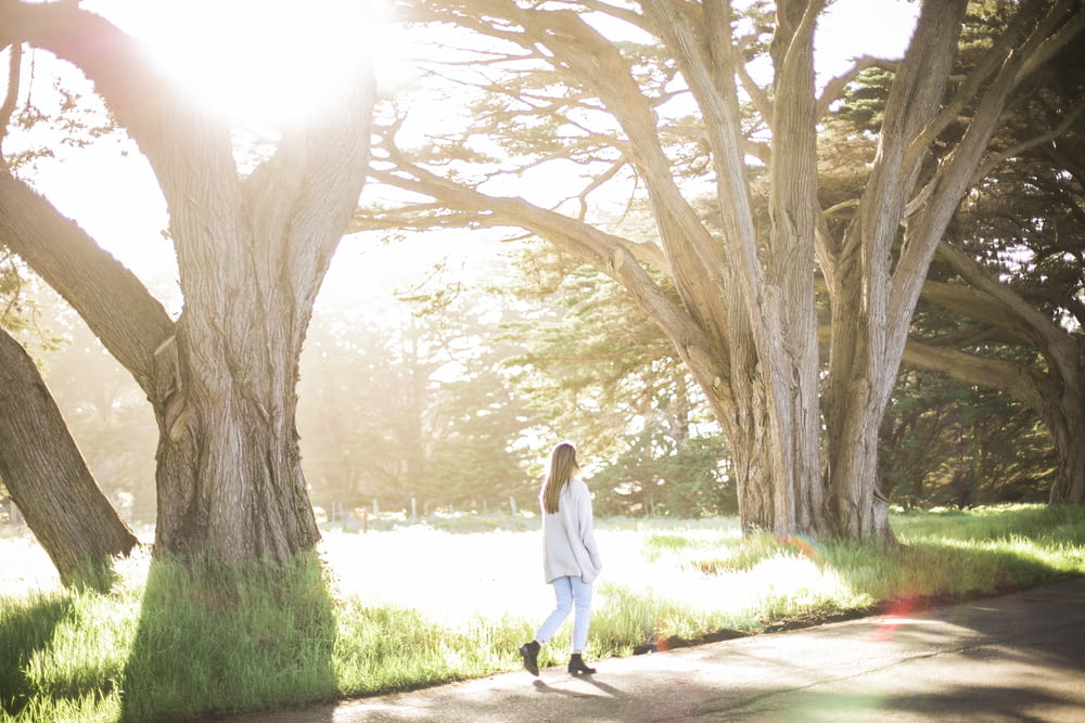 Mujer con camisa blanca de manga larga y pantalones negros caminando por el camino entre los árboles durante el día