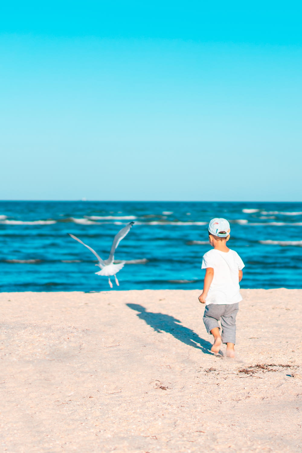menino em camiseta branca e shorts marrons em pé na areia marrom perto do corpo de água