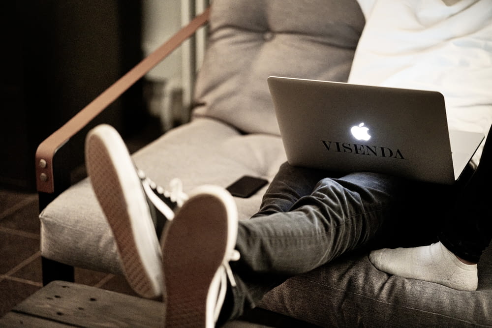 man in white long sleeve shirt and blue denim jeans sitting on brown sofa using macbook