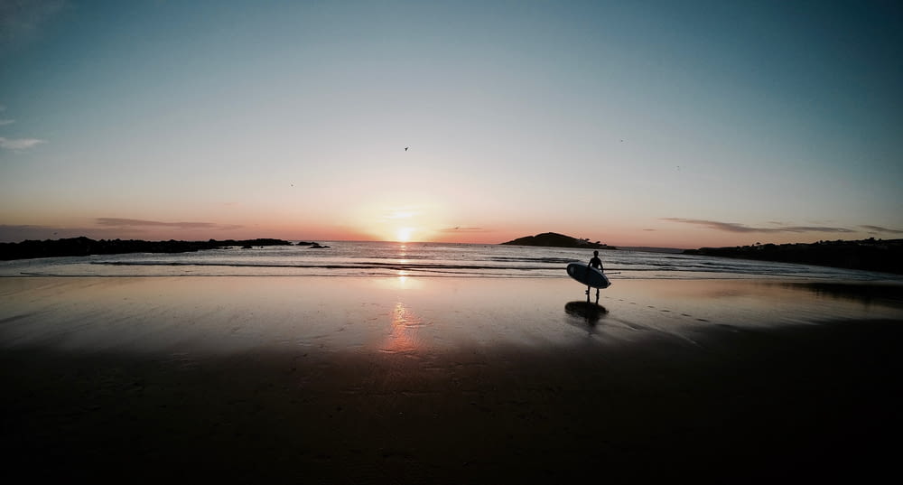silhouette of person carrying surfboard walking on beach during sunset