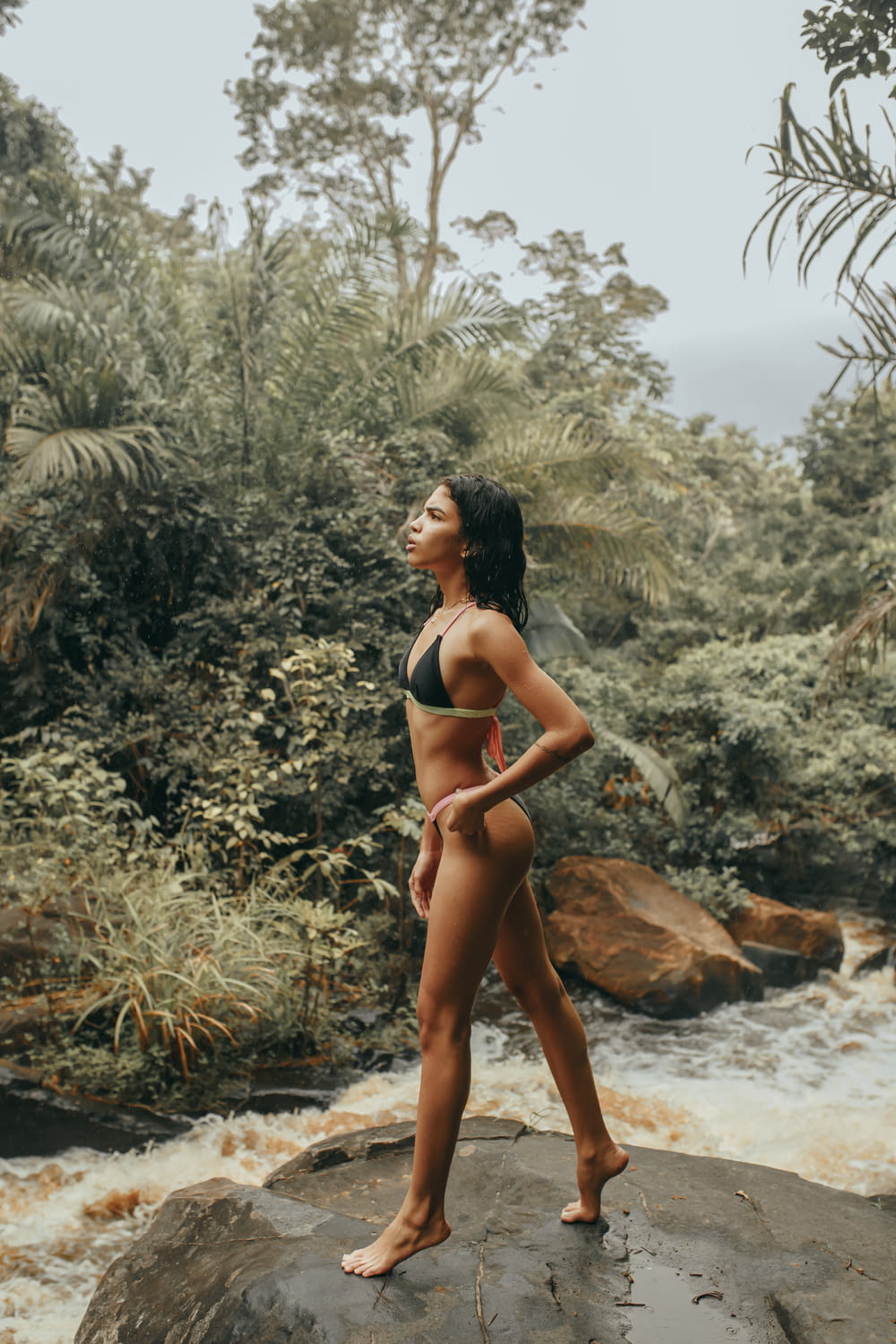woman in black bikini standing on brown rock near river during daytime