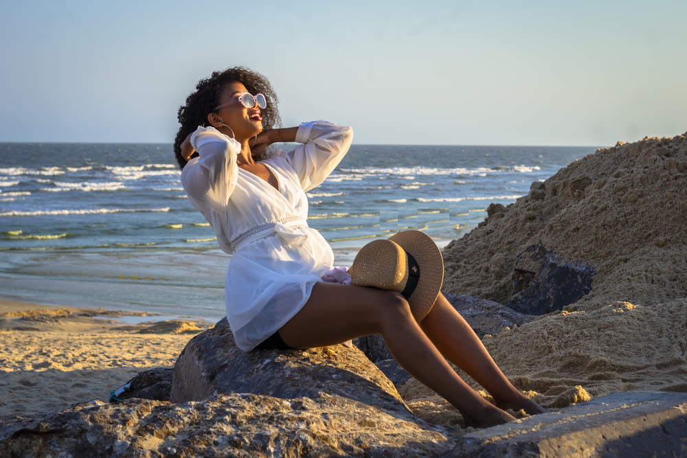 woman in white dress shirt and black skirt sitting on brown rock near body of water