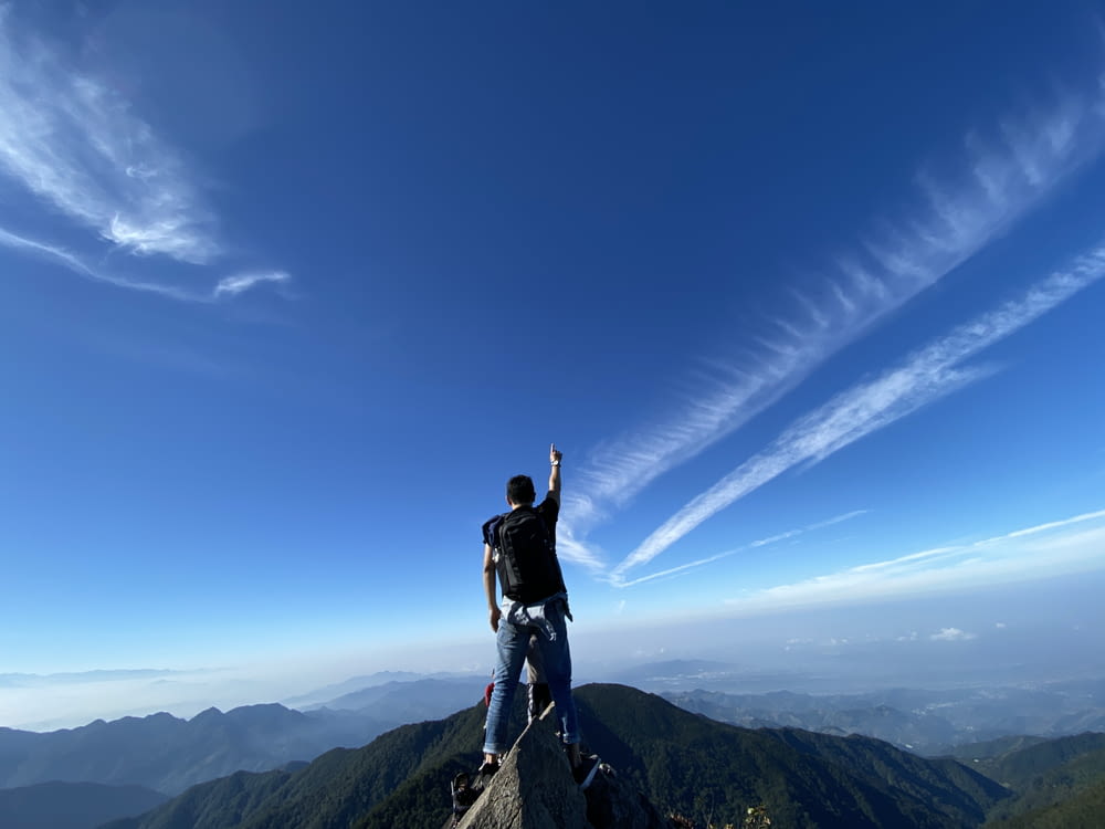 man in black jacket and blue denim jeans standing on gray rock mountain under blue sky