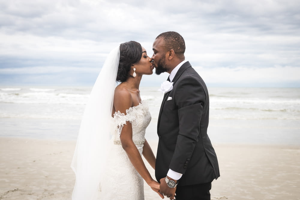 man in black suit kissing woman in white wedding dress on beach during daytime