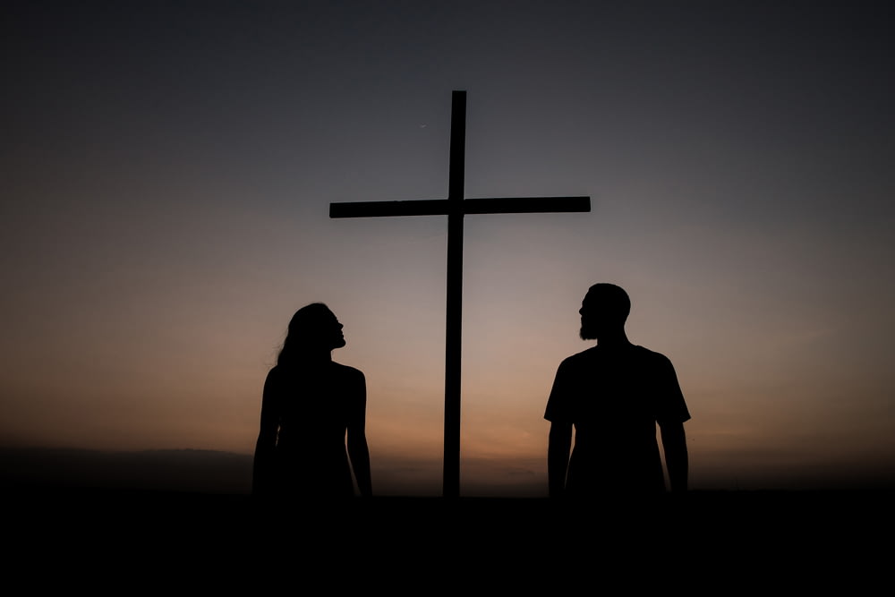 夕暮れ時の十字架の脇に立つ男女のシルエット