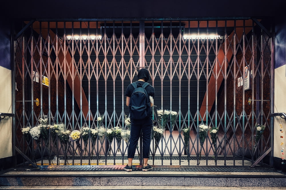 黒い金属製のフェンスの前に立つ黒いジャケットと青いデニムジーンズの男