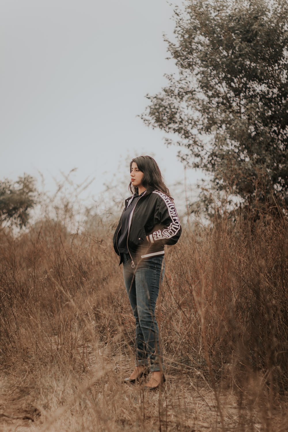 Donna in giacca nera e jeans blu in denim in piedi sul campo di erba marrone durante il giorno
