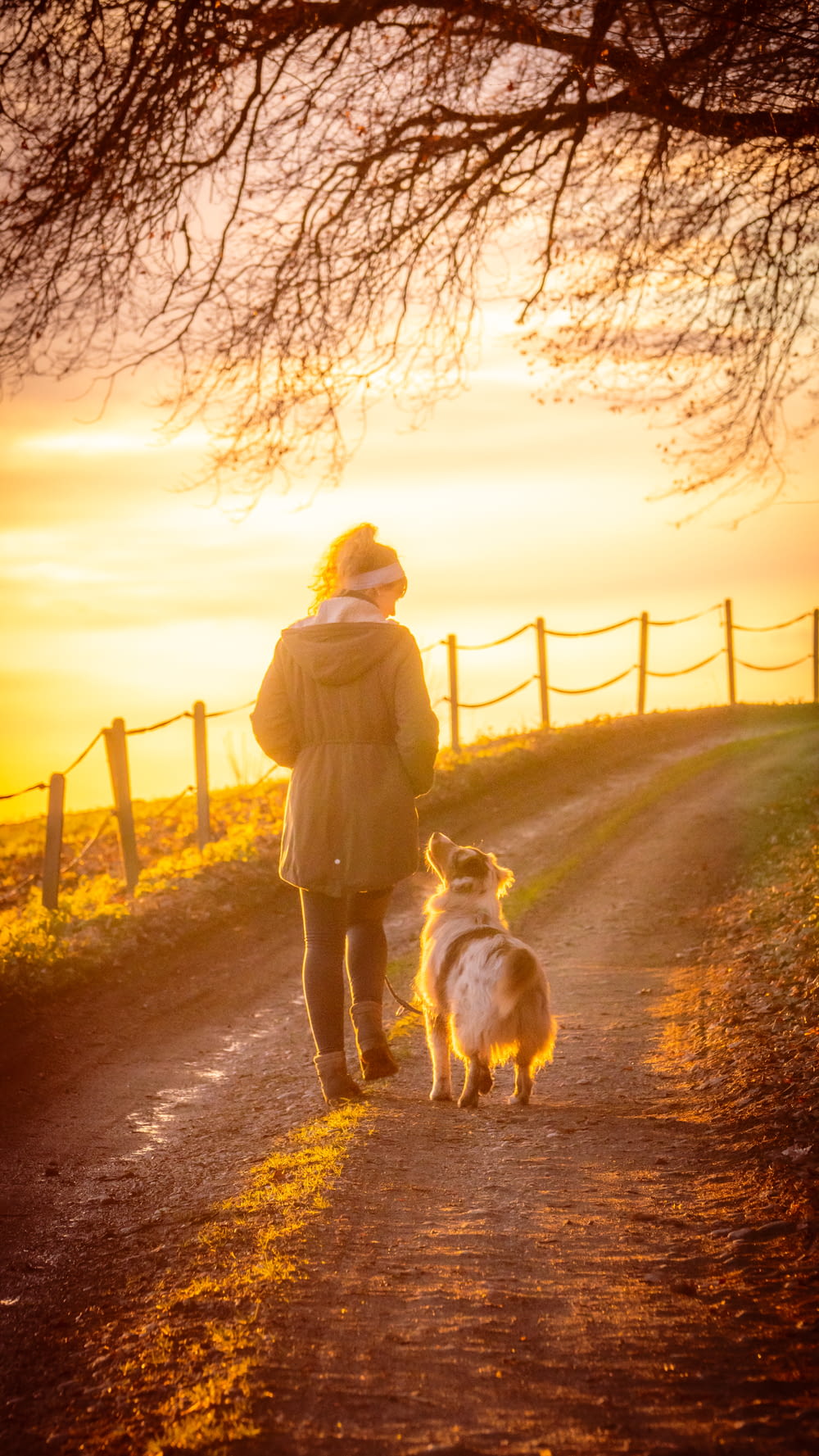 Femme en manteau brun tenant un chien blanc sur un chemin de terre brun au coucher du soleil