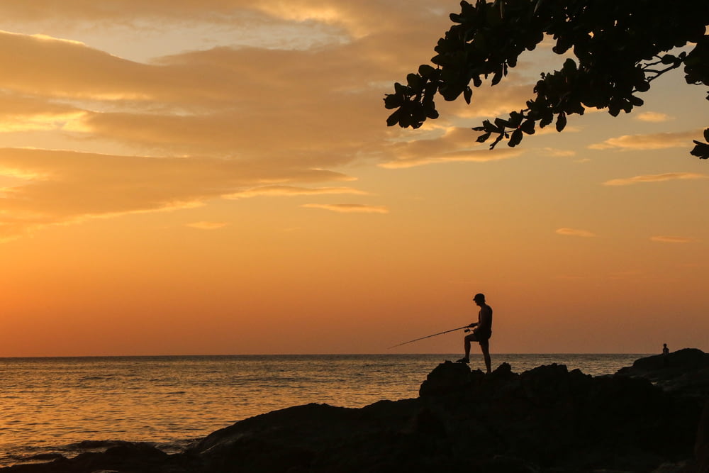 silhouette of man standing on rock near body of water during sunset