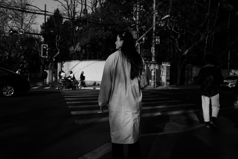 grayscale photo of woman in white coat standing on pedestrian lane